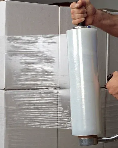 A hand holding a roll of stretch film by its handle while wrapping a stack of boxes.